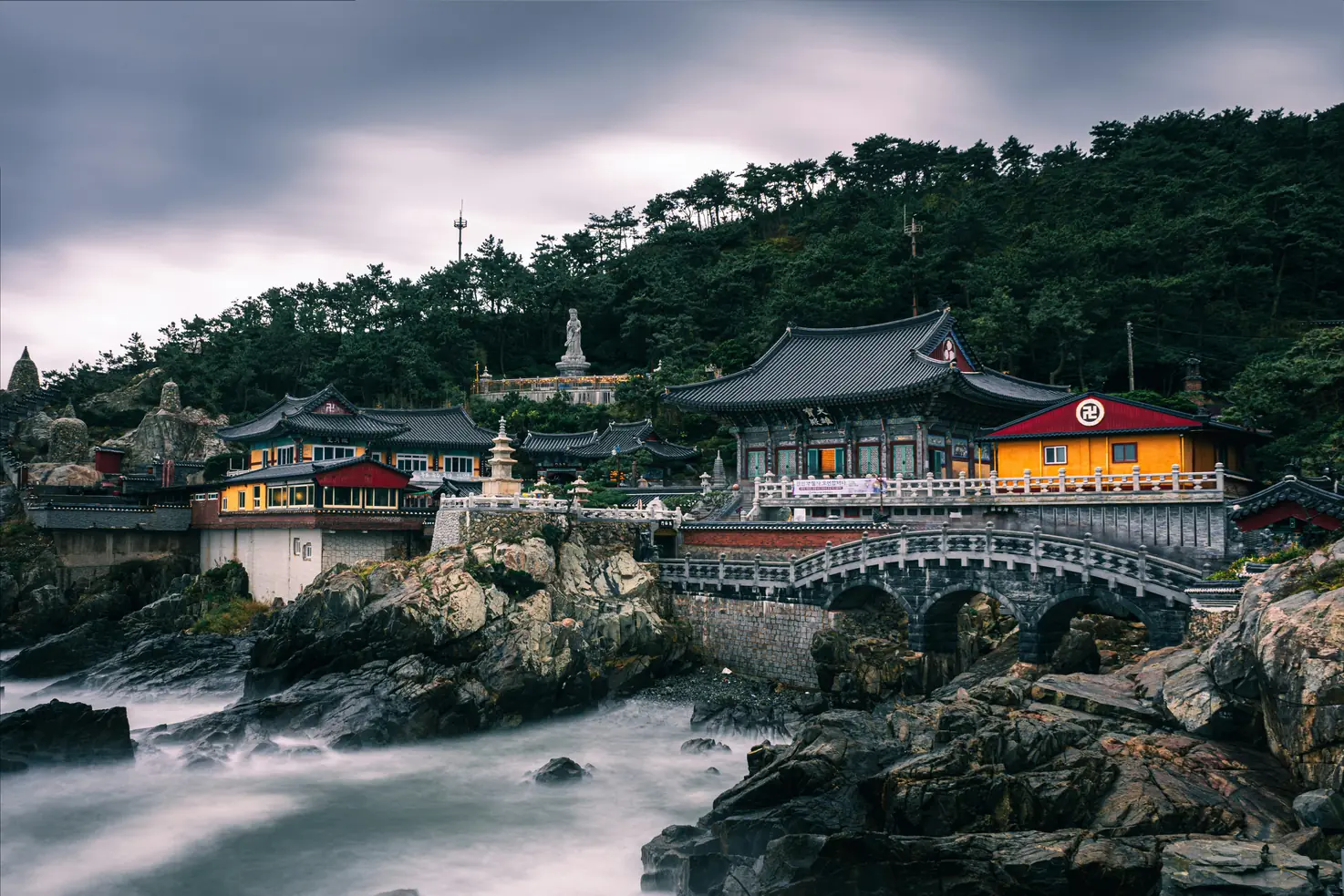 Temple halls and a stone pagoda built on a rocky shoreline above the sea, with an arched bridge below