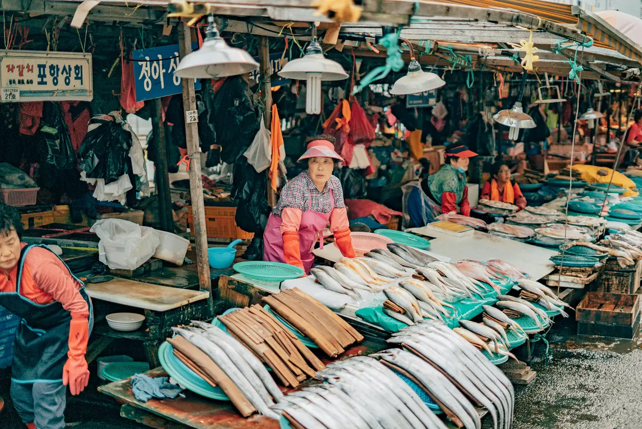 Vendors in aprons behind counters of laid-out fish under a market awning hung with clothing